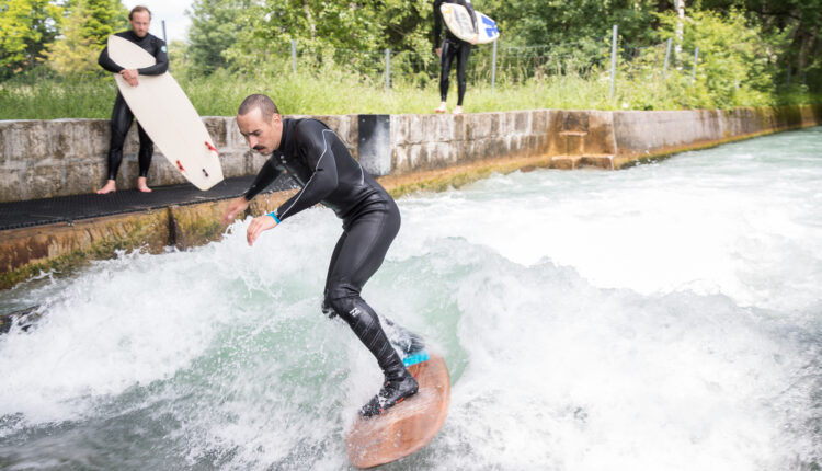 Ein Mann surft auf einer Flusswelle, während zwei Personen mit Surfbrettern vom Betonufer aus zusehen.