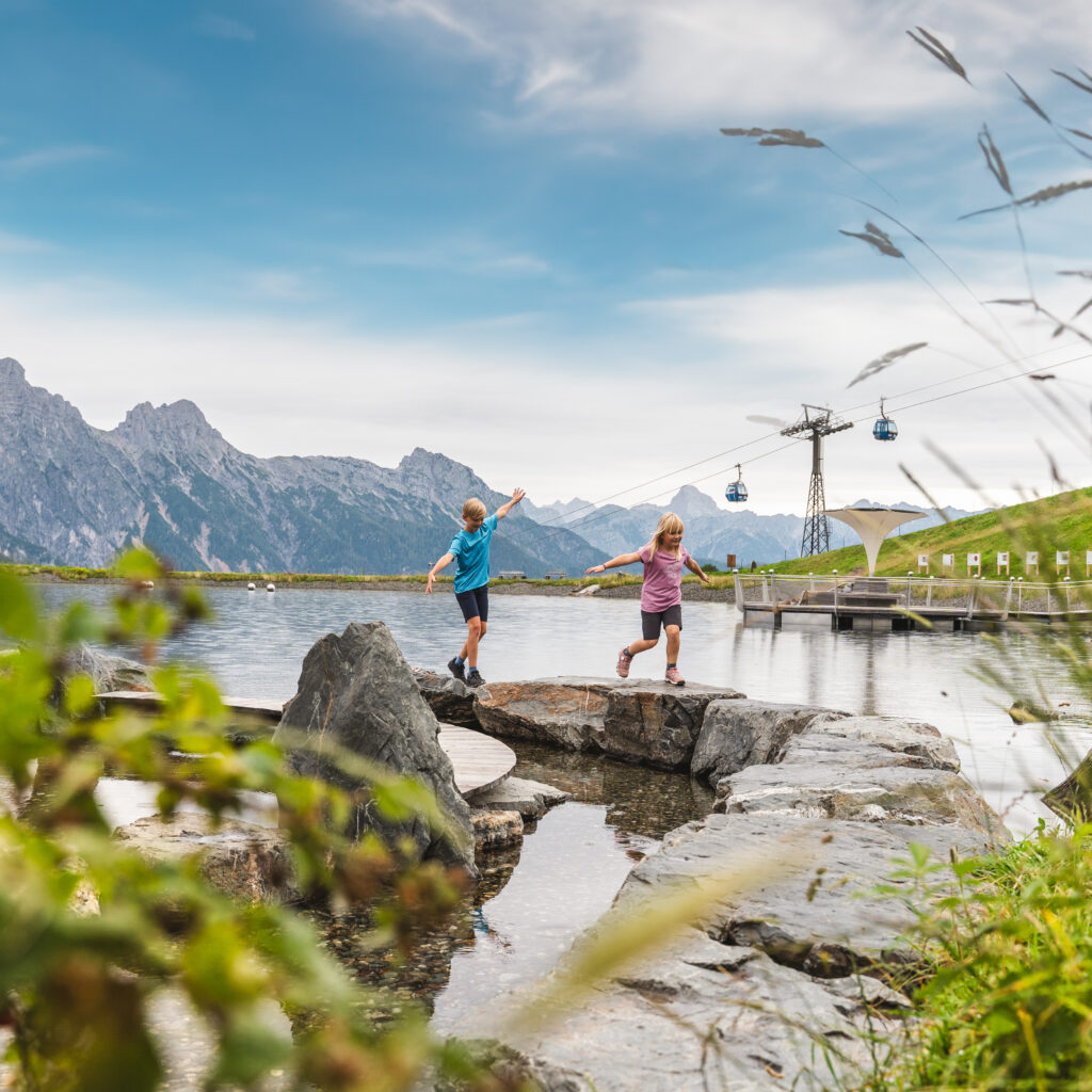 Zwei Kinder spielen auf Felsen neben einem Bergsee, mit Seilbahnen und Bergen im Hintergrund.