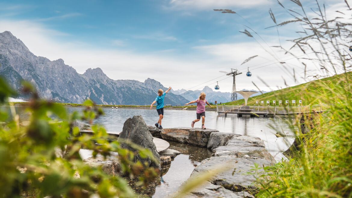 Zwei Kinder spielen auf Felsen neben einem Bergsee, mit Seilbahnen und Bergen im Hintergrund.