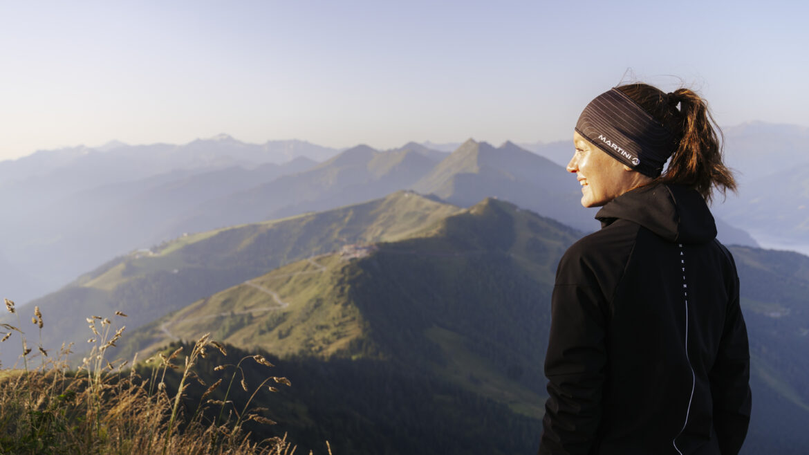 Frau in Sportkleidung steht auf einem Berg, lächelt und schaut auf eine malerische, sonnenbeschienene Berglandschaft.