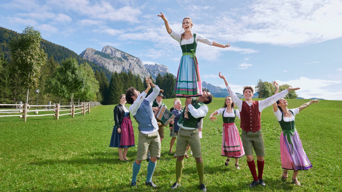 Menschen in traditioneller österreichischer Kleidung führen einen fröhlichen Tanz auf einer Wiese mit Bergen im Hintergrund auf.