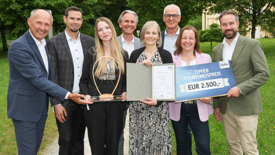 Acht Personen stehen im Freien, lächeln und halten eine Trophäe, eine Urkunde und einen großen Scheck über 2.500 Euro in der Hand.