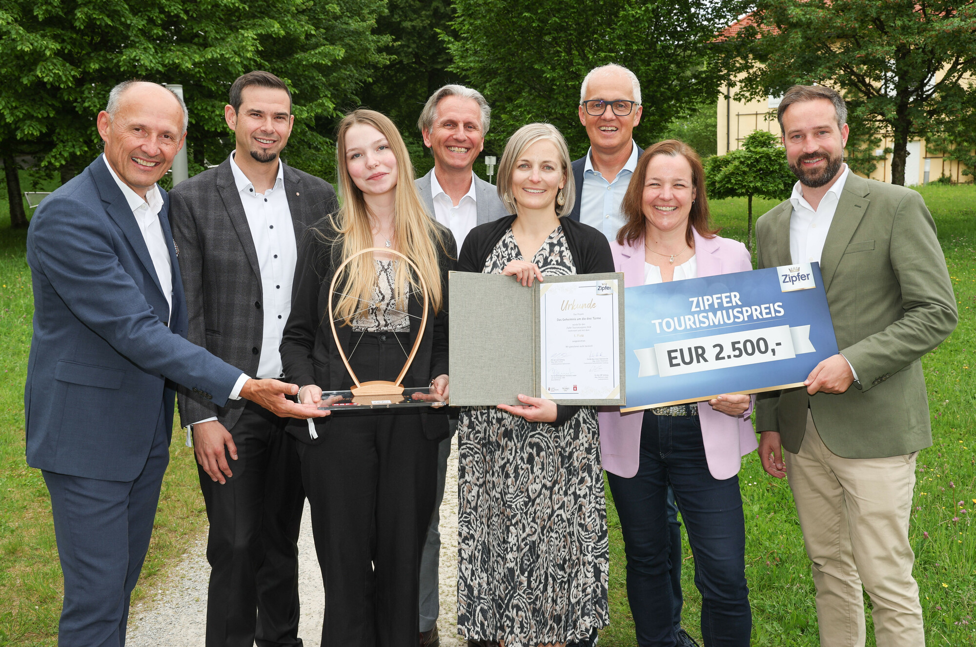 Acht Personen stehen im Freien, lächeln und halten eine Trophäe, eine Urkunde und einen großen Scheck über 2.500 Euro in der Hand.