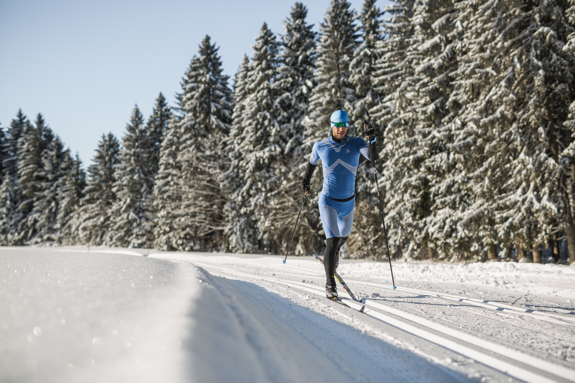 Ein Skifahrer in blauer Ausrüstung fährt Langlaufski auf einer verschneiten Loipe neben schneebedeckten Bäumen unter einem klaren Himmel.