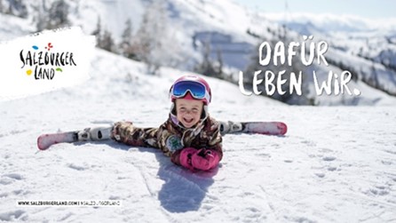 Ein lächelndes Kind in Skiausrüstung macht einen Spagat auf einer verschneiten Piste, mit Bergen im Hintergrund.