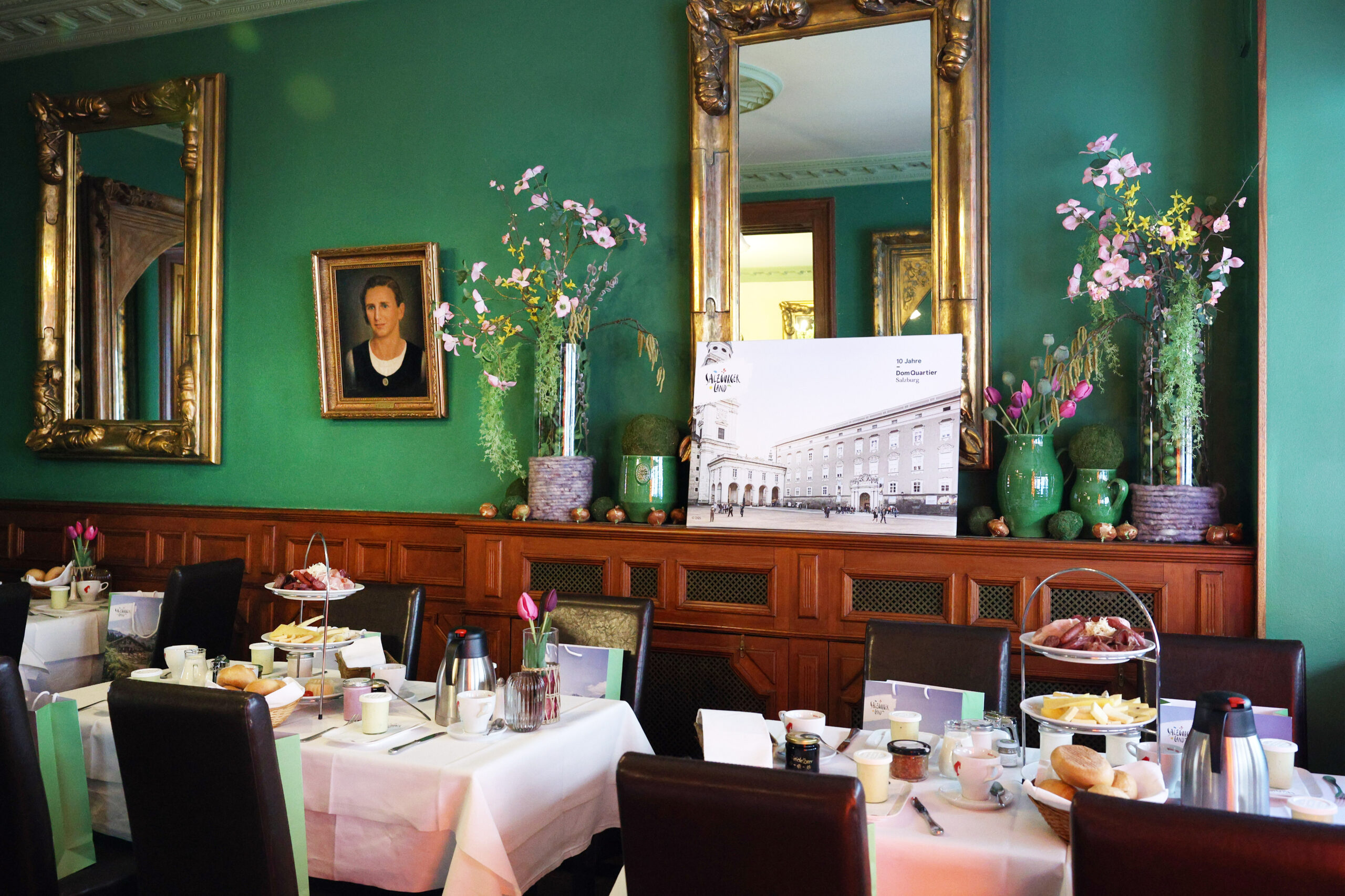 Elegantes Esszimmer mit grünen Wänden, Spiegeln, Blumen und gedeckten Tischen für das Frühstück mit verschiedenen Speisen.