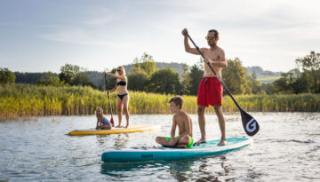 Eine vierköpfige Familie fährt mit dem Paddleboard auf einem ruhigen See, umgeben von Schilf und Bäumen unter einem klaren Himmel.