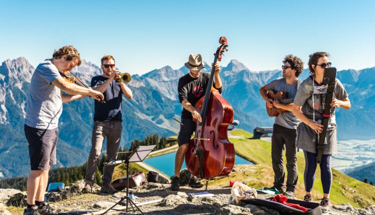 Fünf Musiker spielen Instrumente im Freien auf einem Berg mit blauem Himmel und malerischen Gipfeln im Hintergrund.