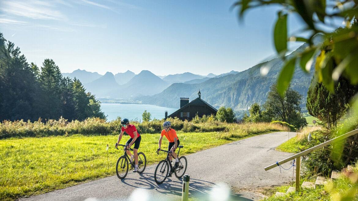Zwei Radfahrer fahren auf einer malerischen Straße an einem See mit Bergen und einer Holzhütte im österreichischen Stil im Hintergrund.