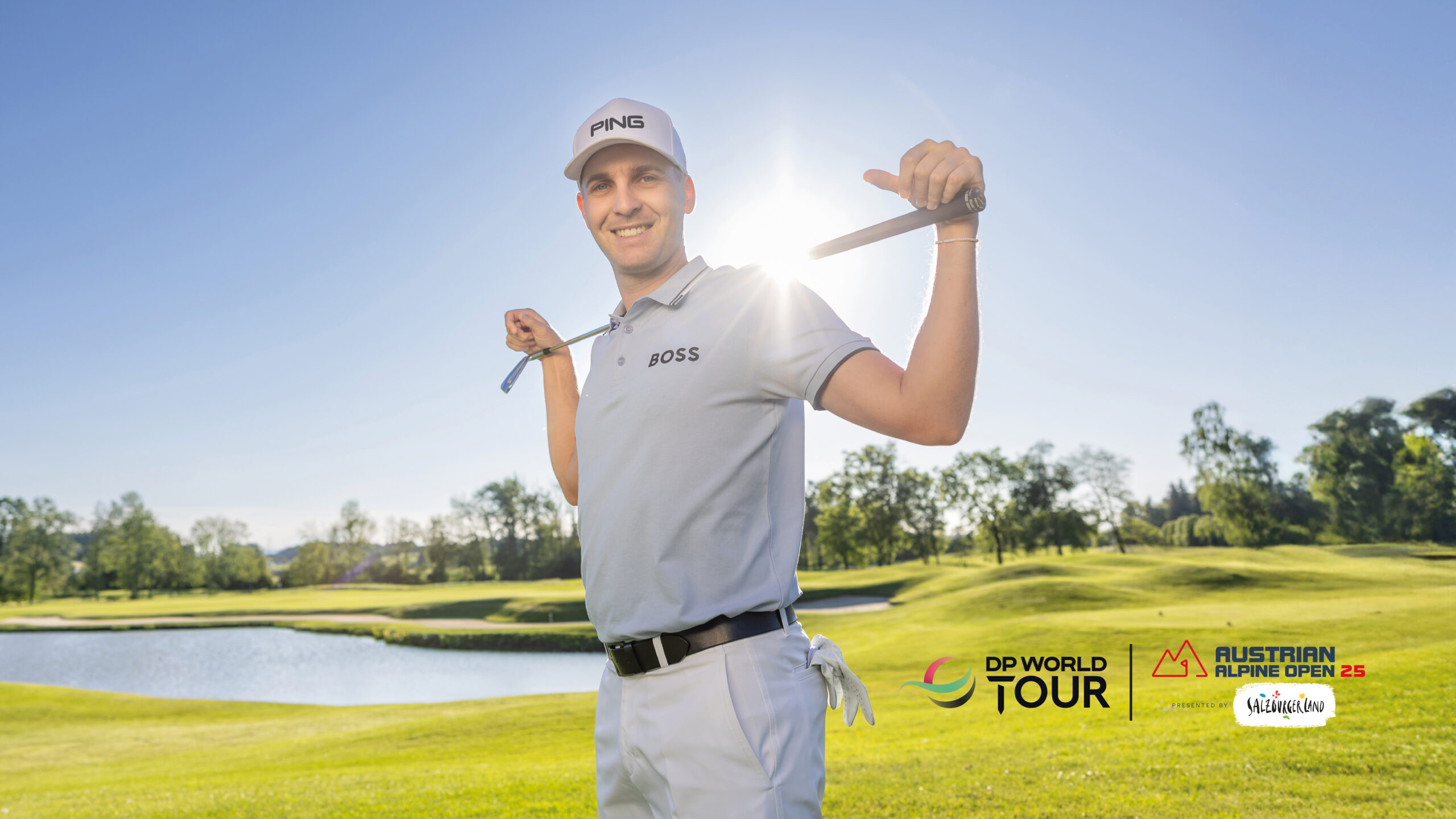 Ein lächelnder Golfer posiert auf einem sonnigen Golfplatz, einen Schläger im Nacken haltend; Turnierlogos in der Ecke.