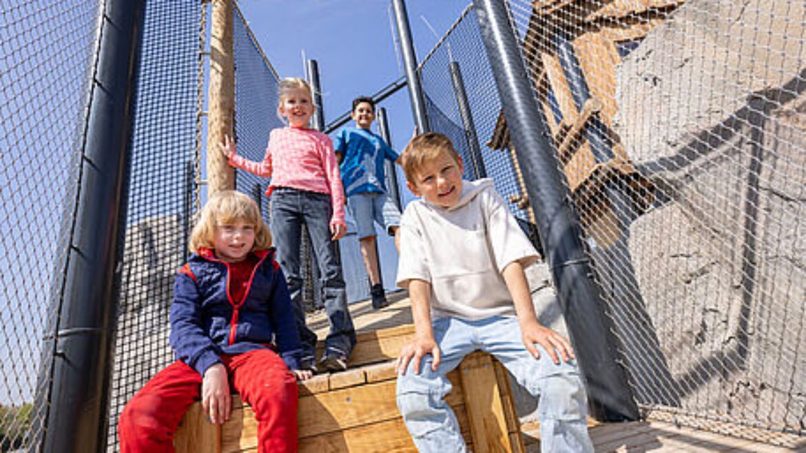 Vier Kinder posieren und lächeln auf den hölzernen Stufen eines von Maschendrahtzäunen umgebenen Spielplatzes.