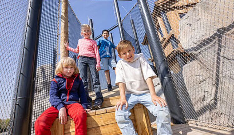 Vier Kinder posieren und lächeln auf den hölzernen Stufen eines von Maschendrahtzäunen umgebenen Spielplatzes.