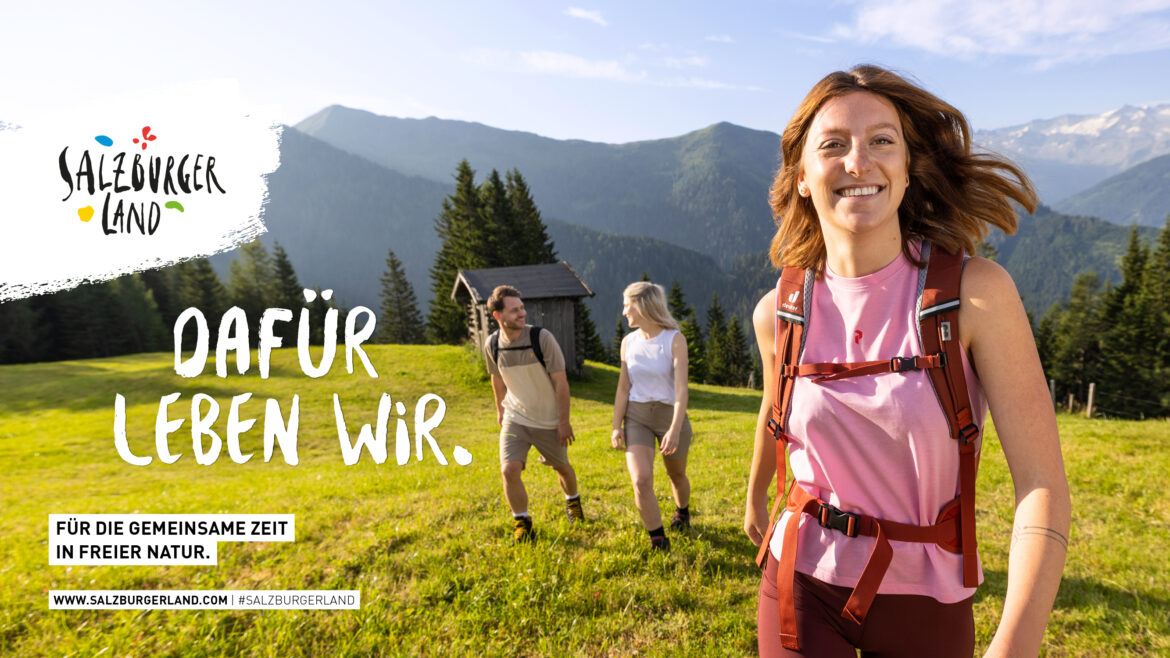 Drei Personen wandern auf einem sonnigen Bergpfad mit grünen Hügeln und blauem Himmel im Hintergrund.