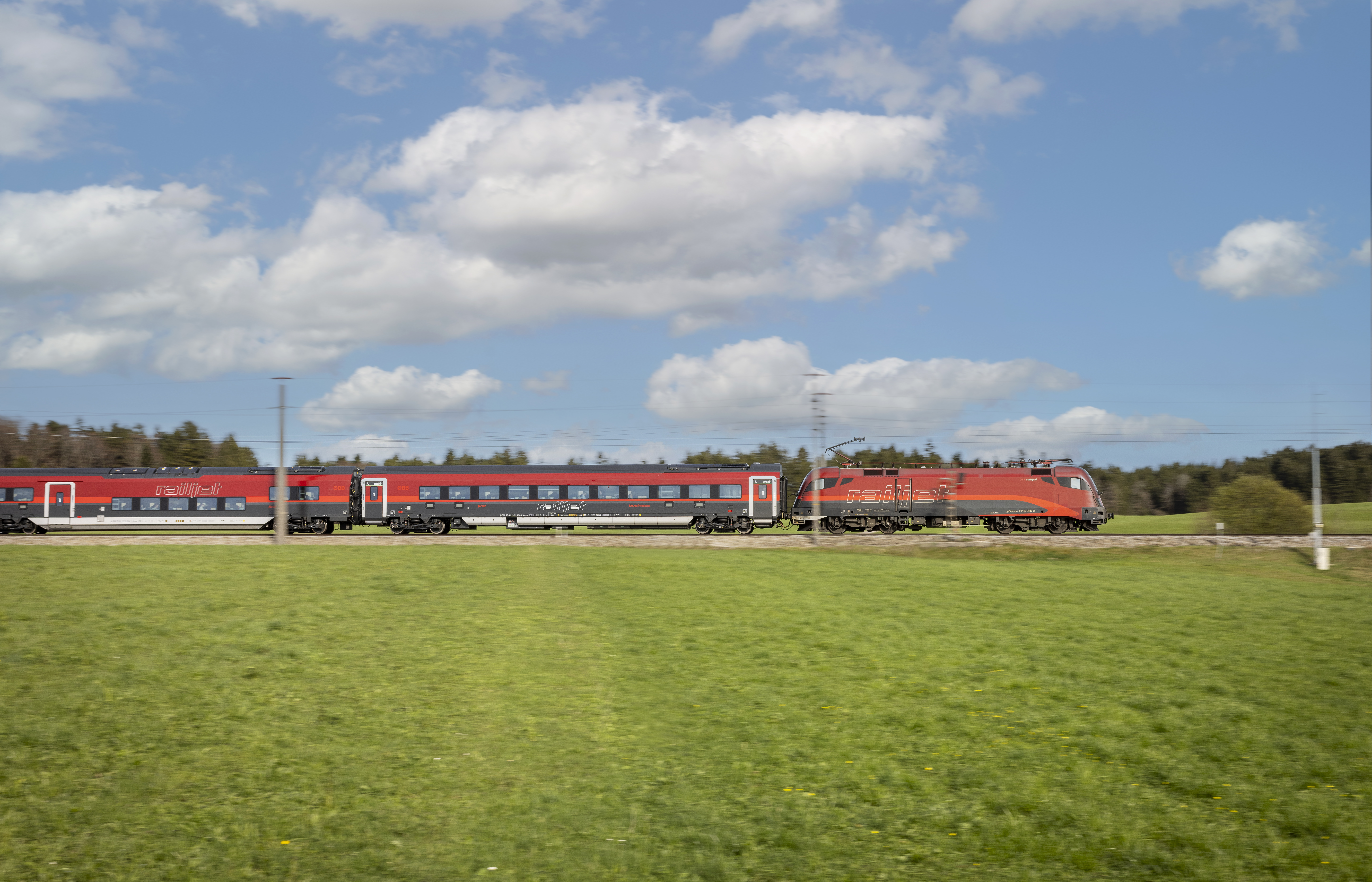 Ein roter Personenzug fährt schnell durch ein grünes Feld unter einem teilweise bewölkten Himmel.