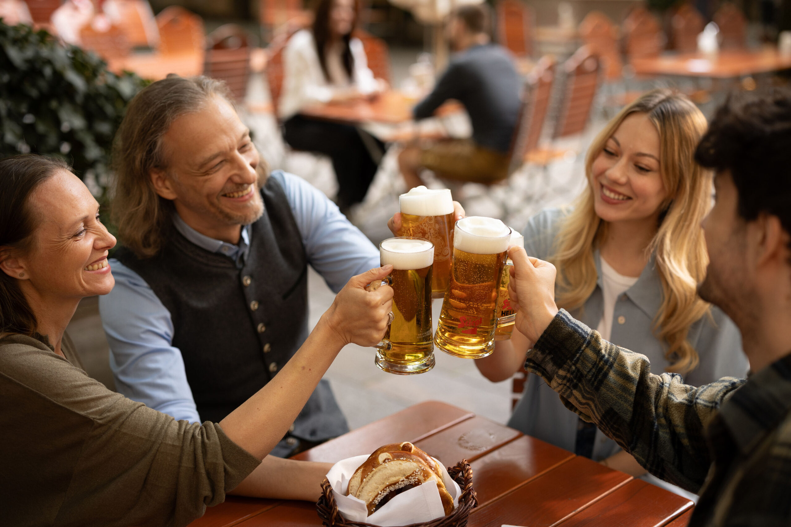 Vier Personen sitzen an einem Tisch im Freien, stoßen mit Biergläsern an und lächeln, während in der Nähe ein Korb mit Essen steht.