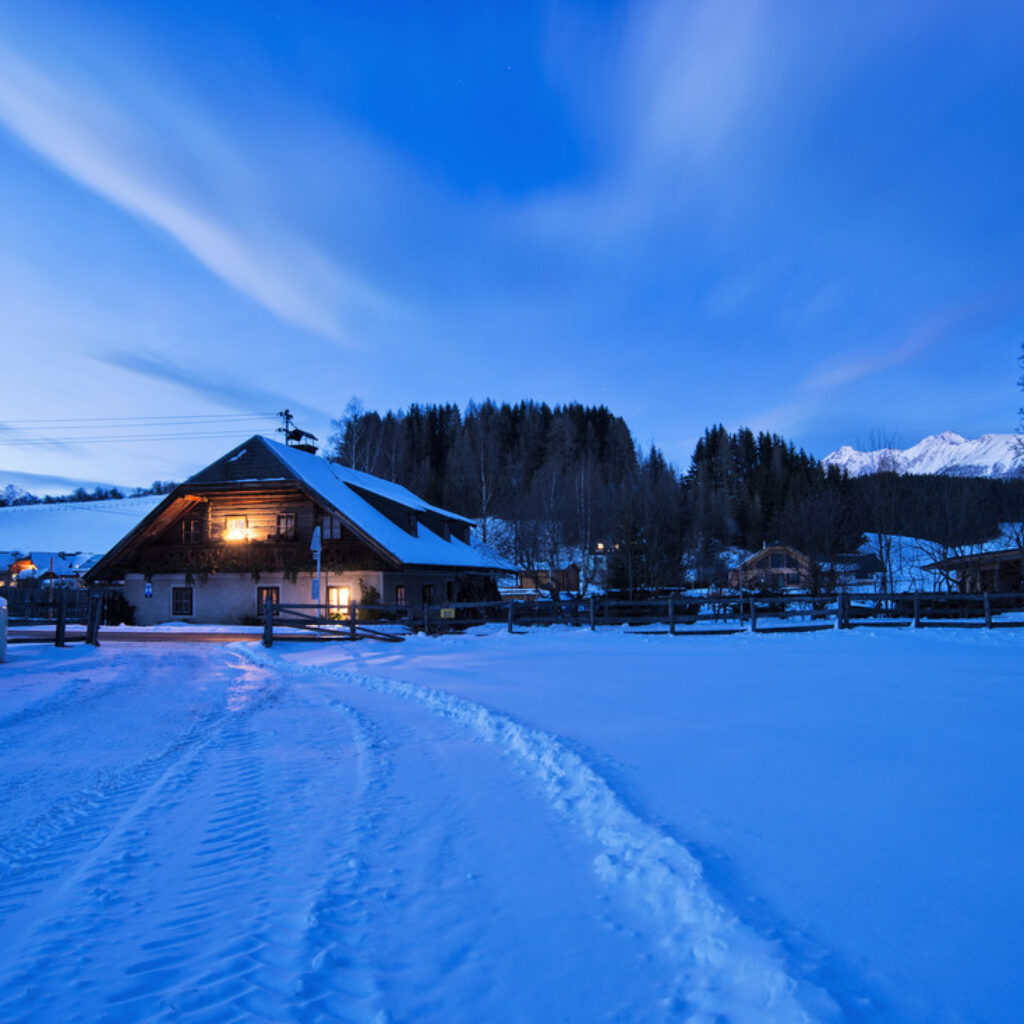 Ein beleuchtetes Bauernhaus bei Nacht, umgeben von einer verschneiten Landschaft mit Bergen im Hintergrund.