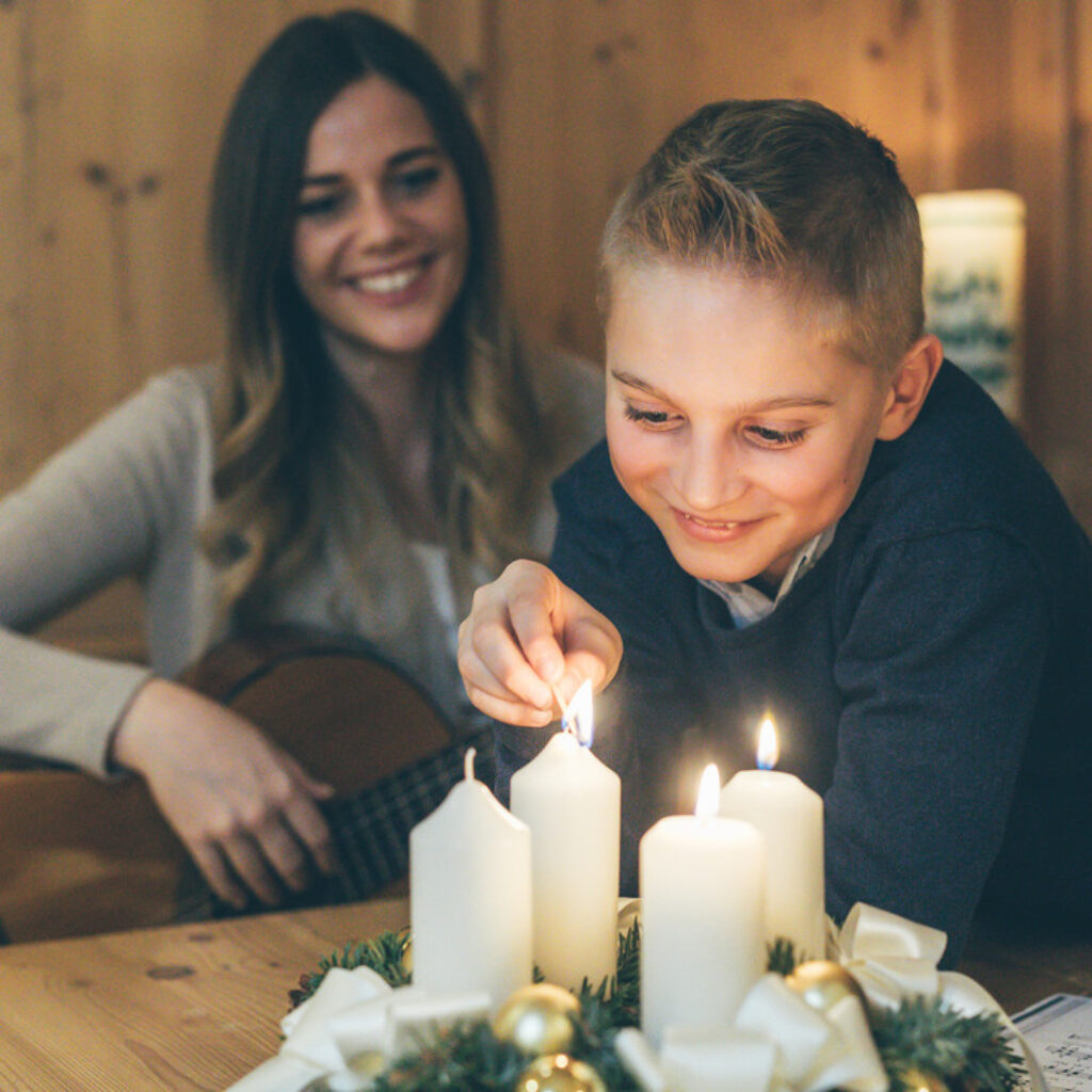 Ein Junge zündet eine Kerze auf einem Adventskranz an, während im Hintergrund eine Frau eine Gitarre in der Hand hält.