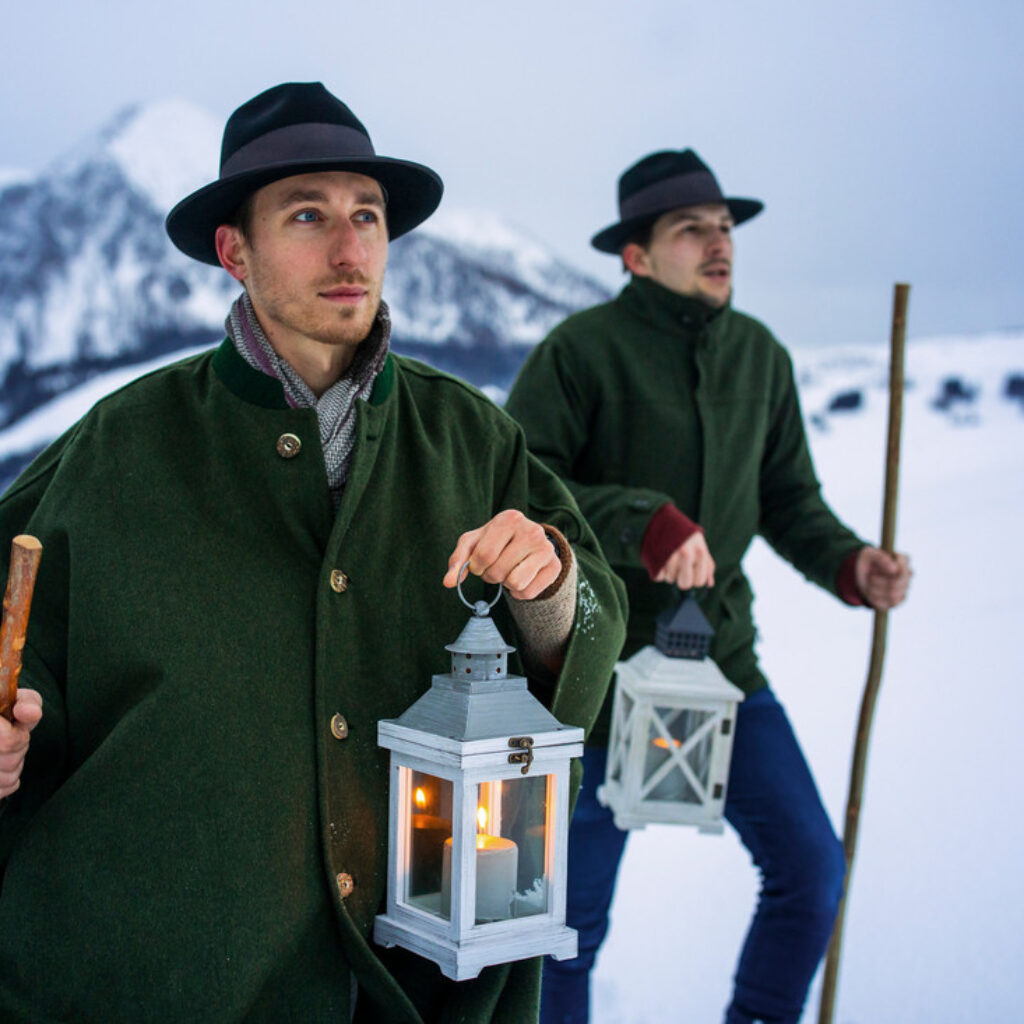 Zwei Männer in traditionellen österreichischen Lodenmänteln und -hüten gehen mit brennenden Laternen durch den Schnee.