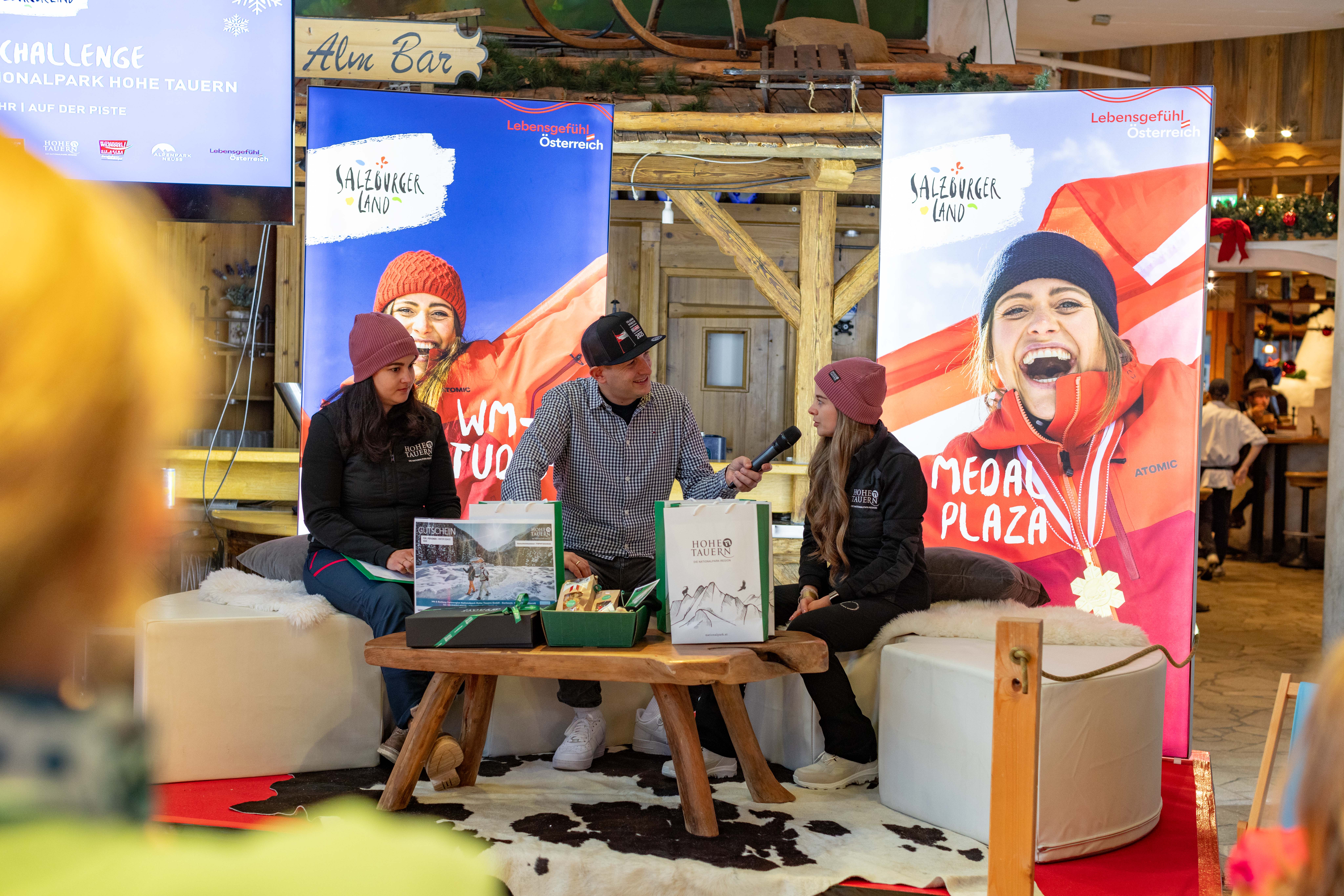 Drei Personen in Winterkleidung unterhalten sich auf einer Bühne mit Bannern und Broschüren zum Thema Skifahren in einem gemütlichen Innenbereich.