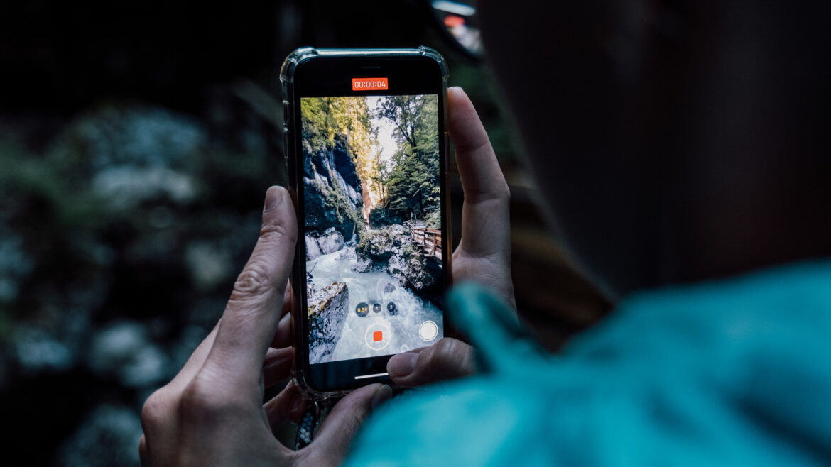 Eine Person hält ein Telefon in der Hand und nimmt ein Video eines Waldbachs und eines Wasserfalls auf dem Telefondisplay auf.