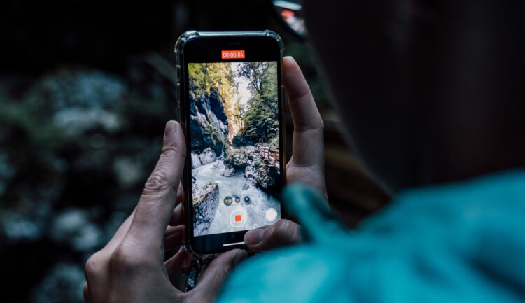 Eine Person hält ein Telefon in der Hand und nimmt ein Video eines Waldbachs und eines Wasserfalls auf dem Telefondisplay auf.