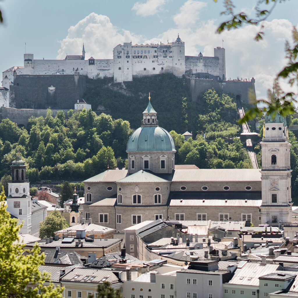 Blick auf den Salzburger Dom mit der Festung Hohensalzburg auf einer Bergkuppe, eingerahmt von grünen Bäumen und blauem Himmel.