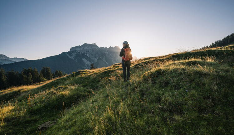 Eine Person wandert bei Sonnenaufgang auf einem grasbewachsenen Hügel, mit Bergen und Bäumen im Hintergrund.