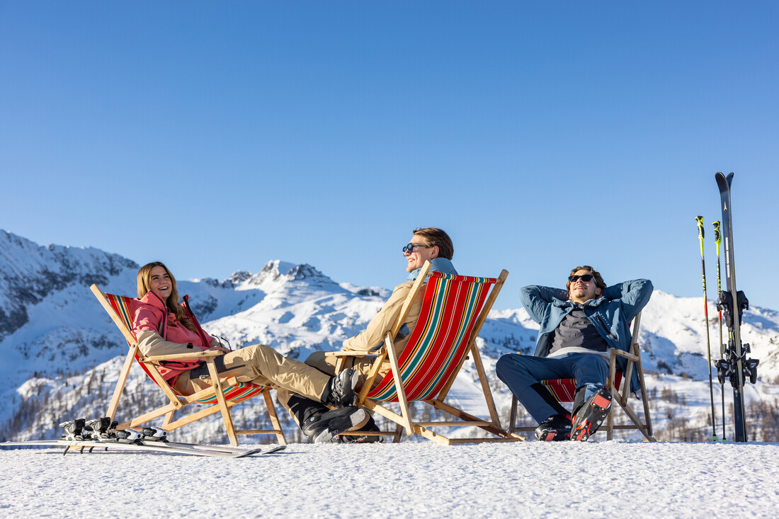 Drei Personen entspannen sich in Liegestühlen auf einem verschneiten Berg mit Skiern in der Nähe unter einem klaren blauen Himmel.