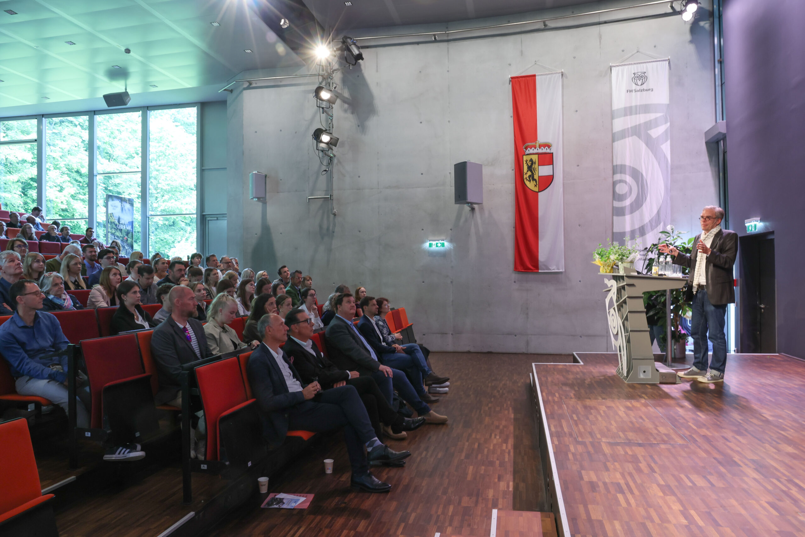 Ein Redner spricht zu einem sitzenden Publikum in einem modernen Hörsaal mit großen Fenstern und einer Flagge an der Wand.
