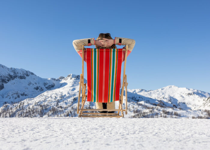 Person, die sich in einem gestreiften Liegestuhl auf verschneitem Boden entspannt, mit Blick auf schneebedeckte Berge unter einem klaren blauen Himmel.