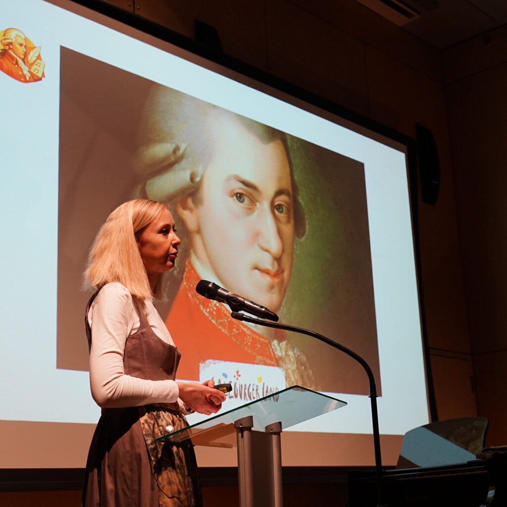 Eine Frau spricht auf einem Podium vor einem großen Bildschirm, der ein Porträt von Mozart zeigt.