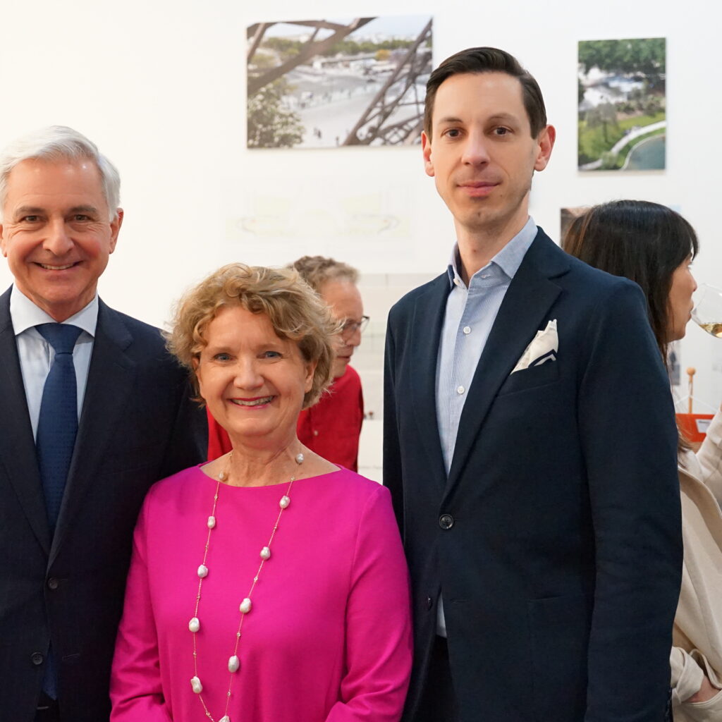 Drei formell gekleidete Personen lächeln bei einer Veranstaltung in einer Kunstgalerie, während sich andere im Hintergrund unter sie mischen.