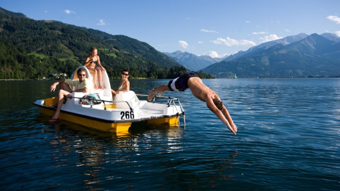 Vier Personen sitzen auf einem Tretboot auf einem See, ein Mann springt ins Wasser, im Hintergrund sind Berge zu sehen.