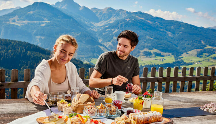 Ein lächelndes Paar genießt das Frühstück im Freien mit Bergen und grünen Tälern im Hintergrund.