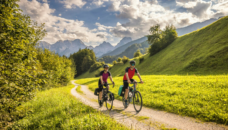 Zwei Radfahrer fahren auf einem gewundenen Weg durch eine grüne Landschaft mit Bergen im Hintergrund.