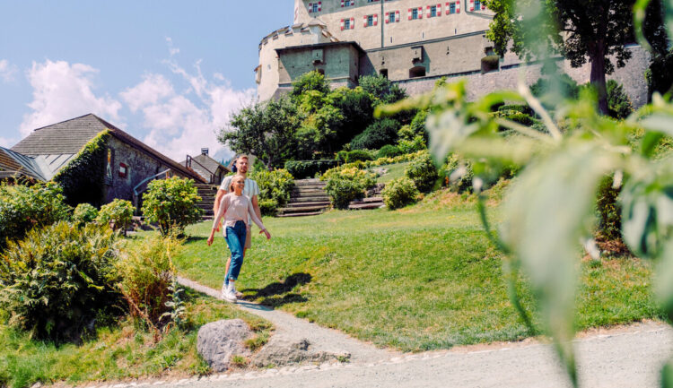 Ein Paar geht an einem sonnigen Tag einen Gartenweg hinauf in Richtung eines großen historischen Gebäudes.