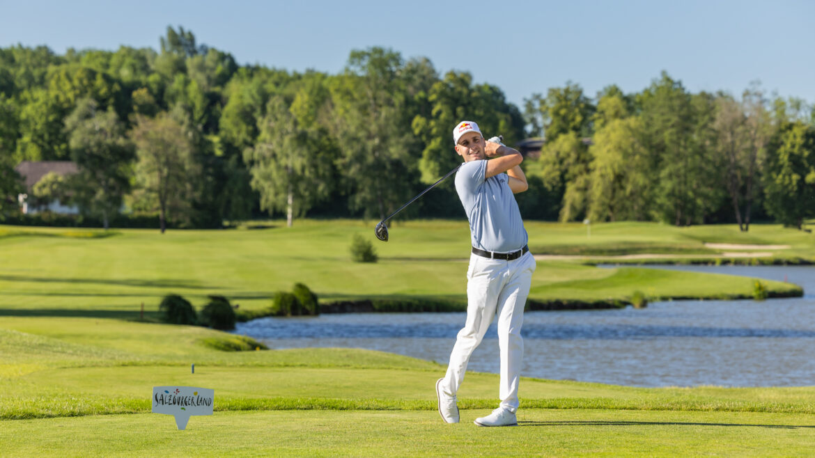 Ein Golfer in weißer Kleidung schwingt einen Schläger auf einem grünen Platz in der Nähe eines Teiches unter einem klaren blauen Himmel.