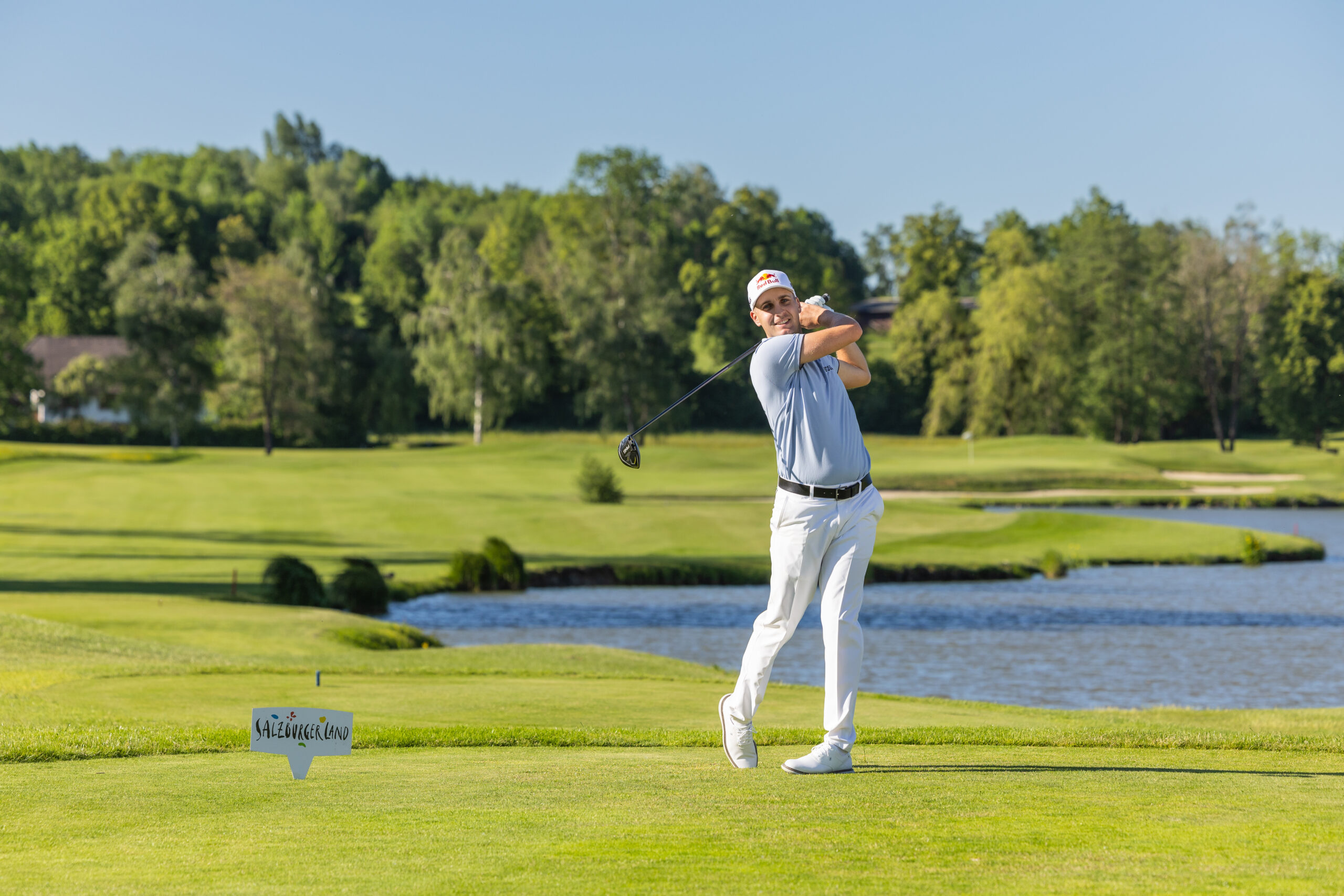 Ein Golfer in weißer Kleidung schwingt einen Schläger auf einem grünen Platz in der Nähe eines Teiches unter einem klaren blauen Himmel.