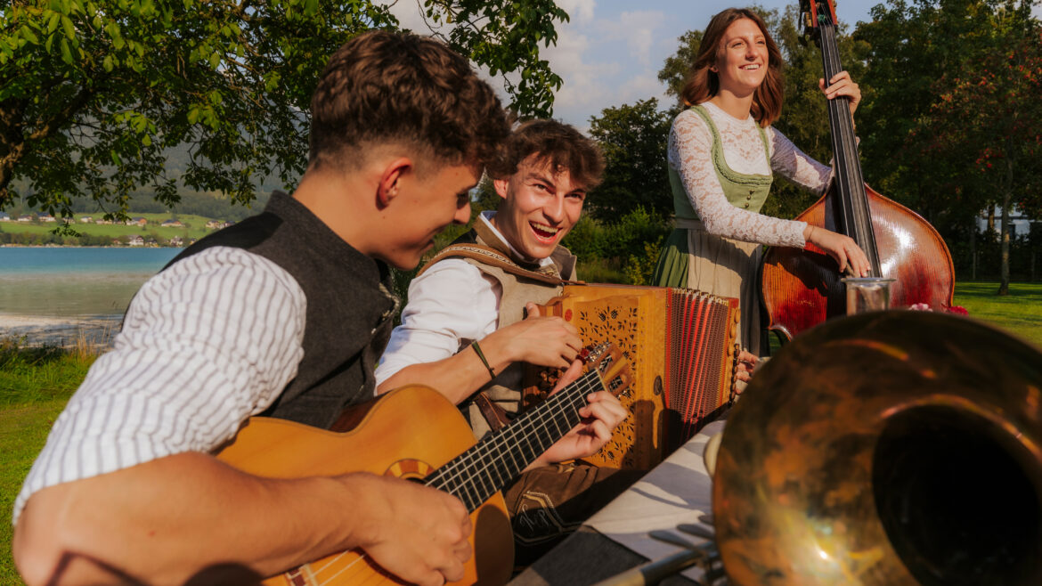 Drei junge Musiker spielen Gitarre, Akkordeon und Kontrabass an einem sonnigen Tag im Freien, lächeln und haben Spaß an der Musik.