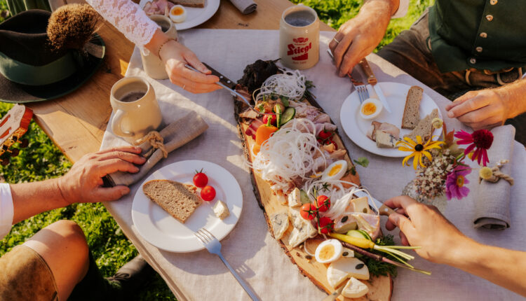 Menschen in traditioneller österreichischer Kleidung teilen sich im Freien eine rustikale Holzplatte mit Fleisch, Eiern, Brot und Gemüse.