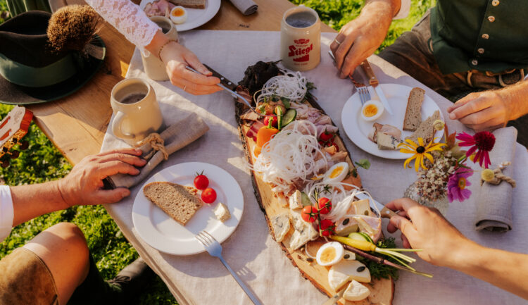 Menschen in traditioneller österreichischer Kleidung teilen sich im Freien eine rustikale Holzplatte mit Fleisch, Eiern, Brot und Gemüse.