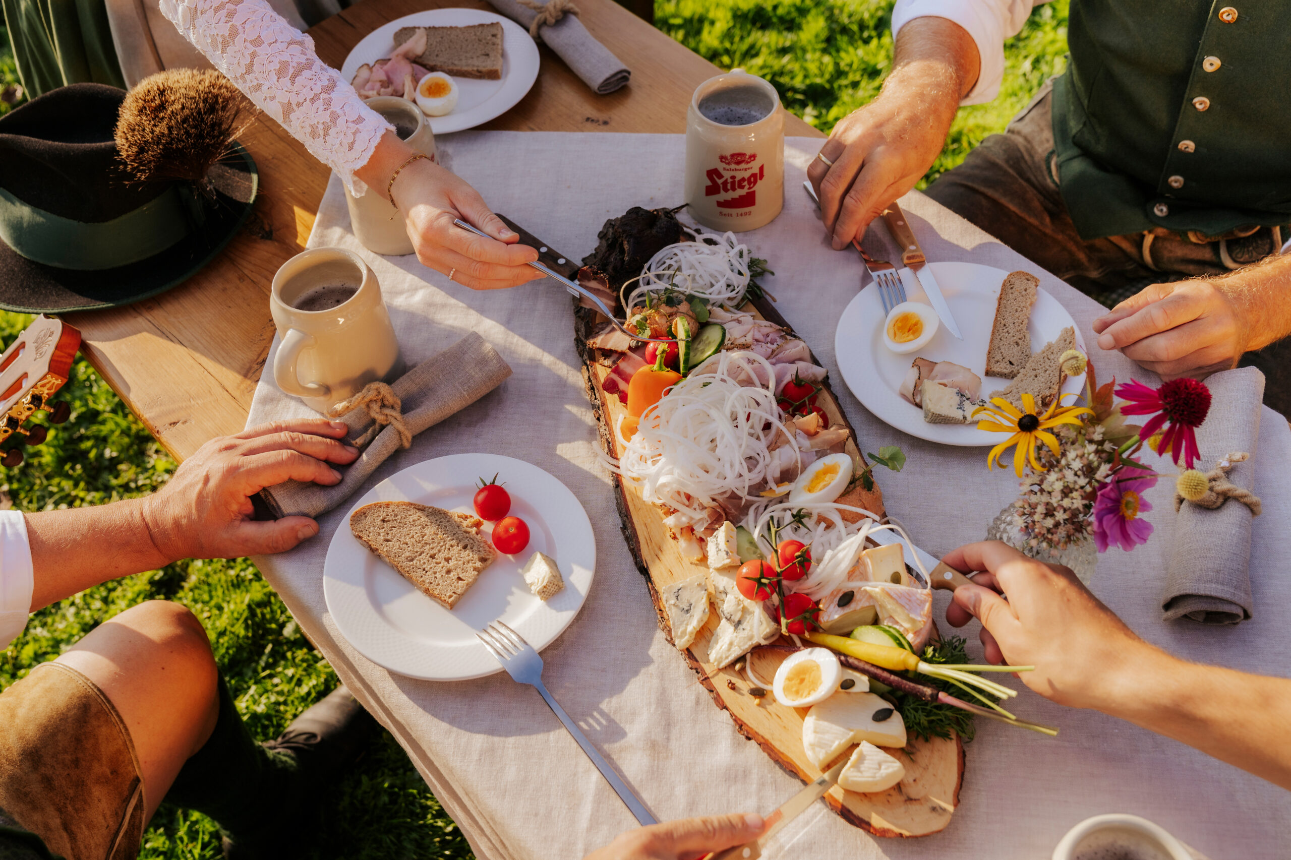 Menschen in traditioneller österreichischer Kleidung teilen sich im Freien eine rustikale Holzplatte mit Fleisch, Eiern, Brot und Gemüse.