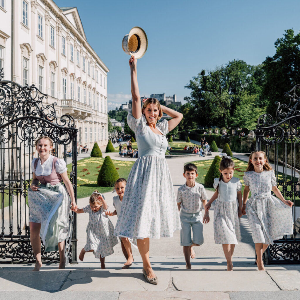 Eine Frau und sechs Kinder in passender Kleidung posieren an einem sonnigen Tag fröhlich vor einem verzierten Gartentor.