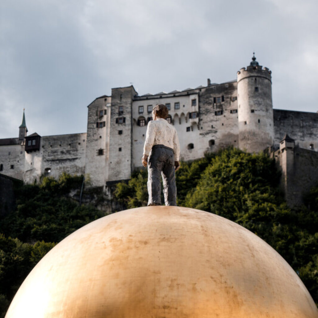 Die Statue einer Person steht auf einer großen goldenen Kugel mit einer Burg auf einem Hügel im Hintergrund.