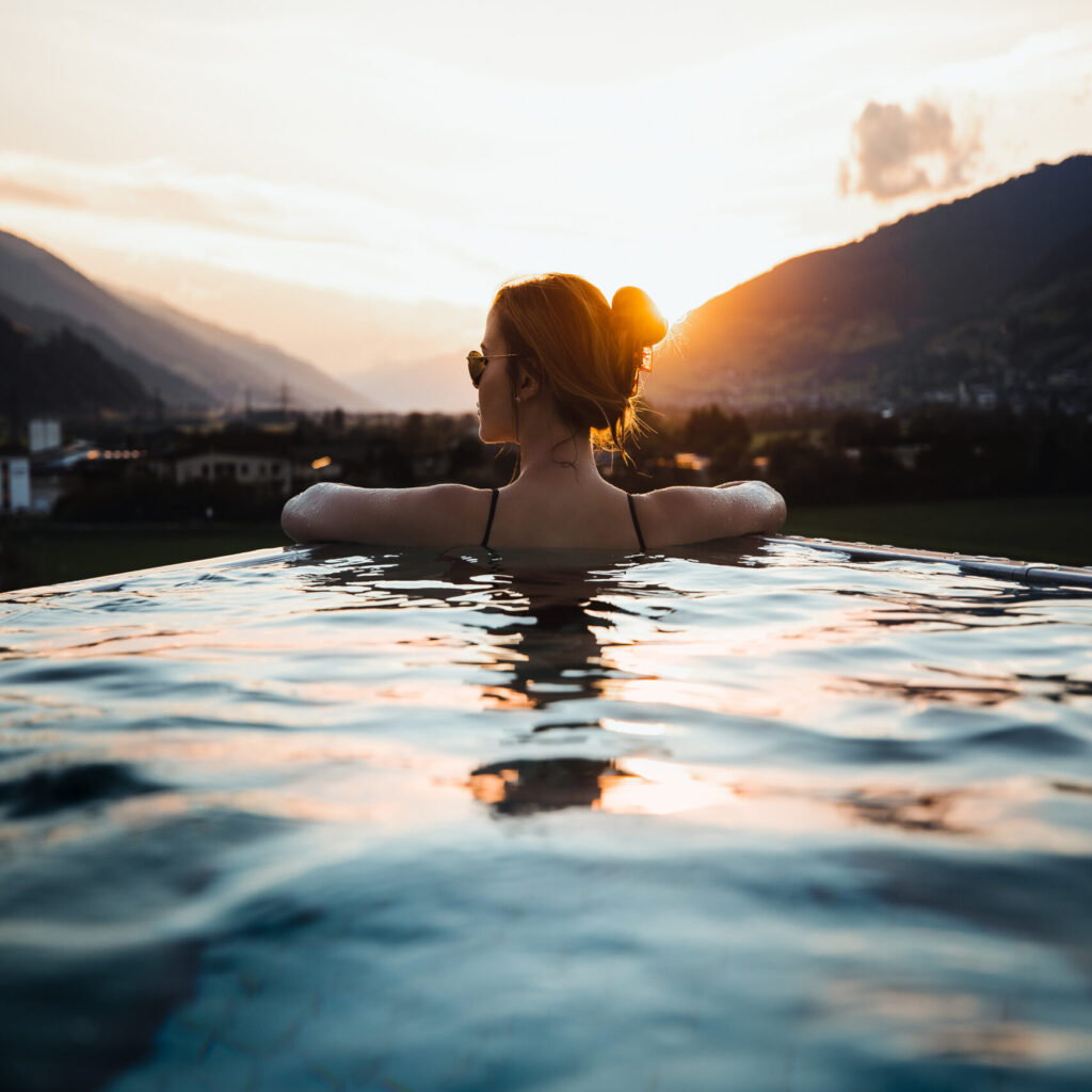 Frau in einem Pool bei Sonnenuntergang, mit Blick auf die Berge in der Ferne mit Sonnenlicht reflektiert auf dem Wasser.