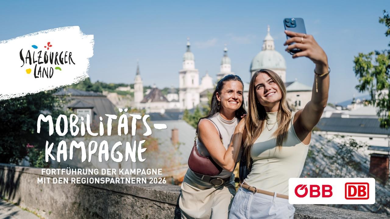 Zwei Frauen lächeln und machen ein Selfie im Freien mit einer Stadtlandschaft und Kuppelgebäuden im Hintergrund.