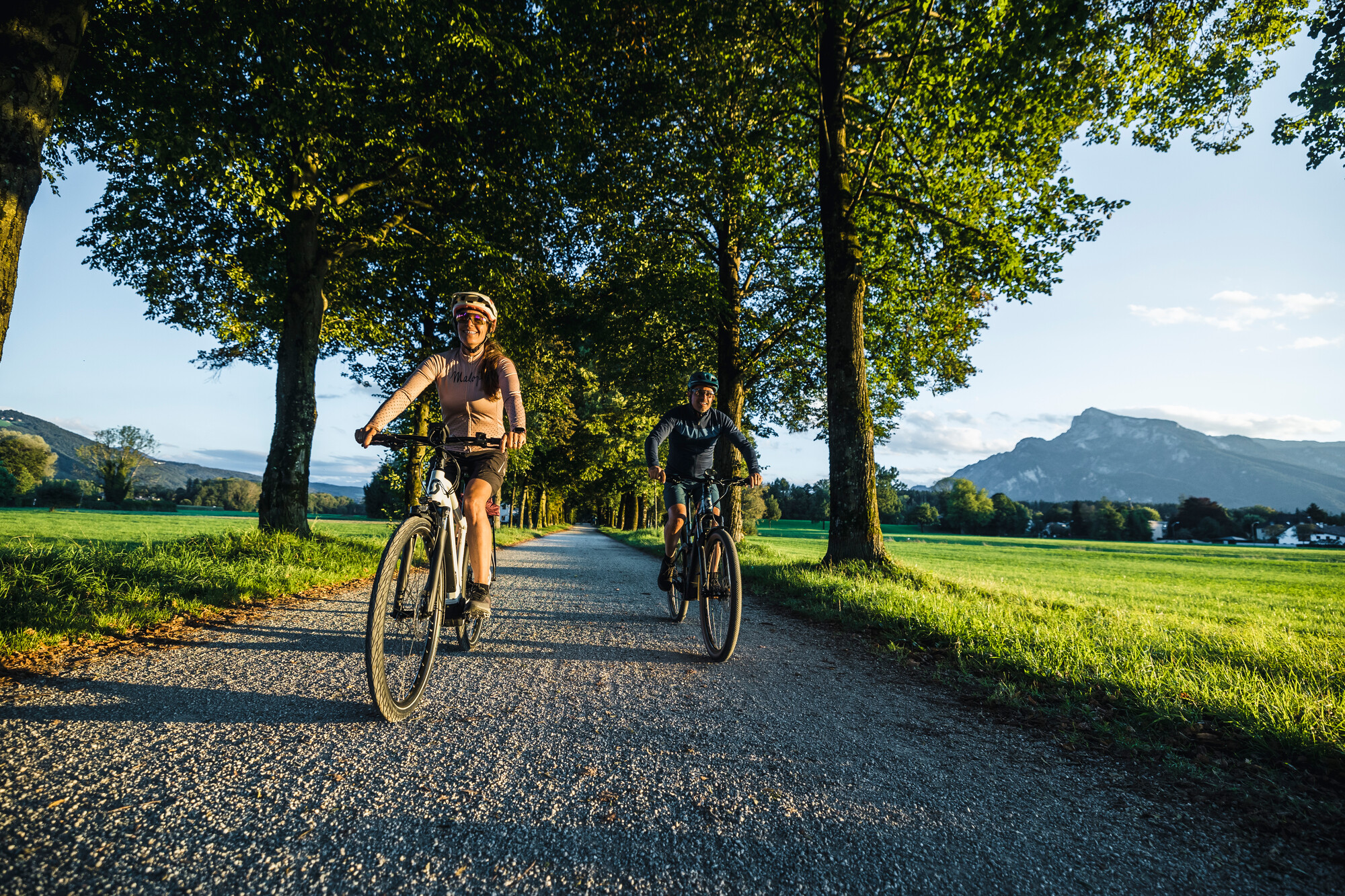 Zwei Personen fahren mit dem Fahrrad auf einem von Bäumen gesäumten Weg auf dem Lande mit Bergen im Hintergrund.