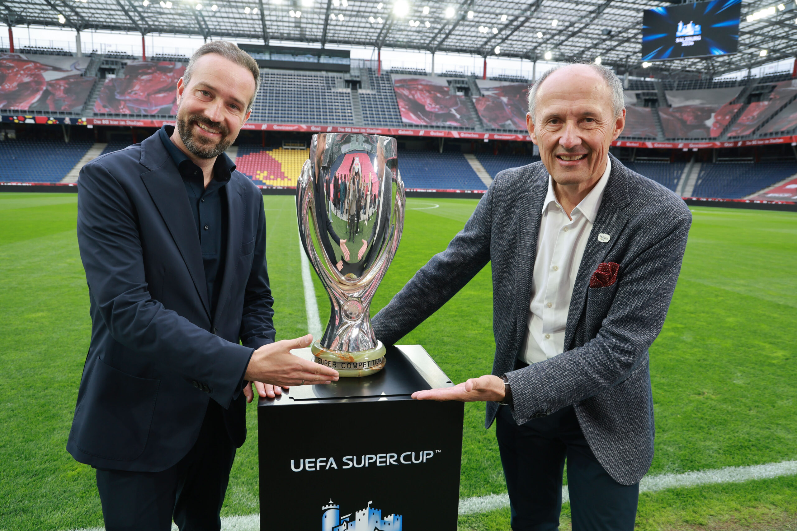 Zwei Männer in Anzügen lächeln, während sie die Trophäe des UEFA-Superpokals auf einem Stadionfeld halten.