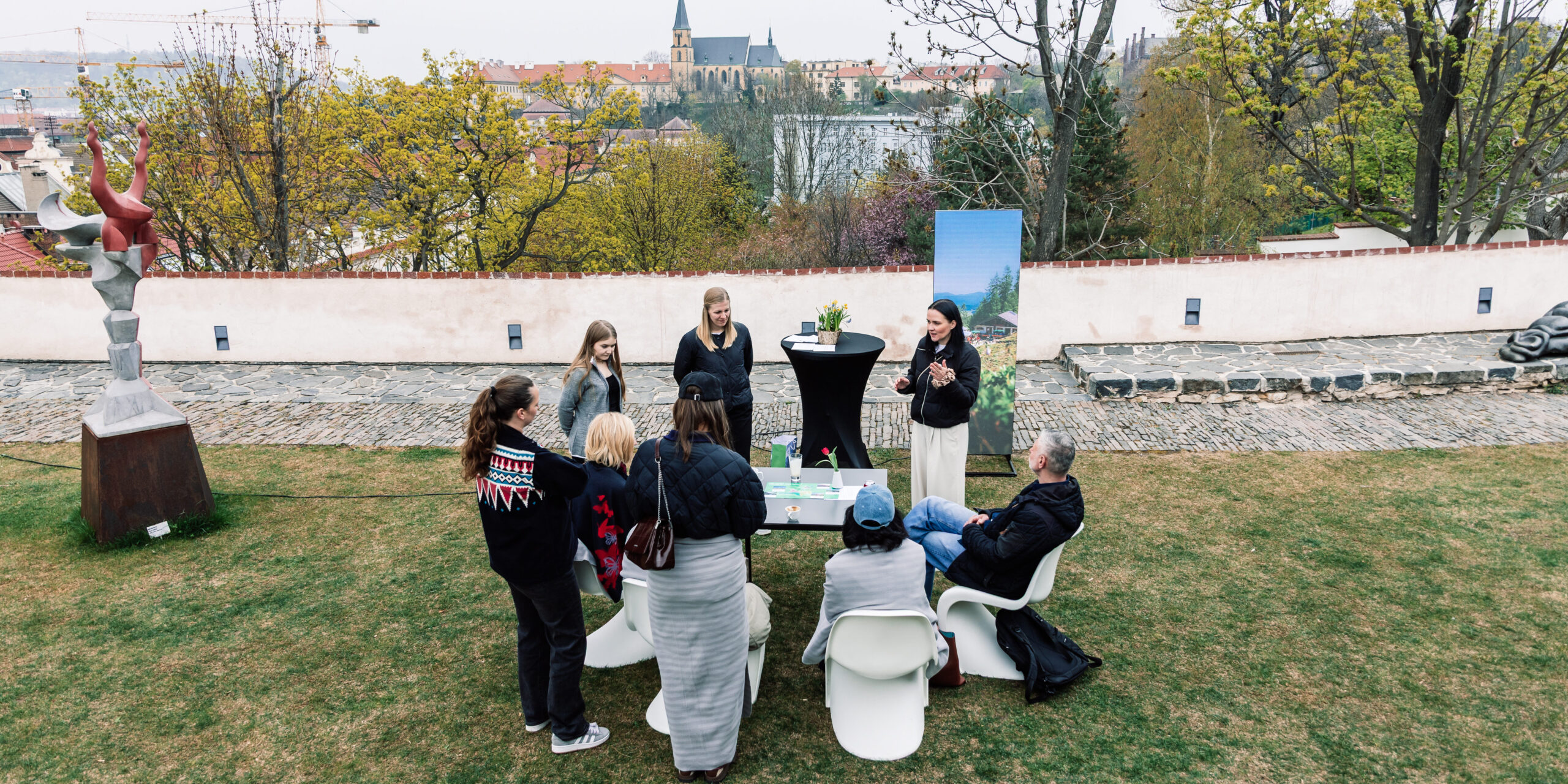 Eine Gruppe von Menschen versammelt sich im Freien um einen Tisch in einem Garten mit Blick auf die Stadt im Hintergrund und unterhält sich.