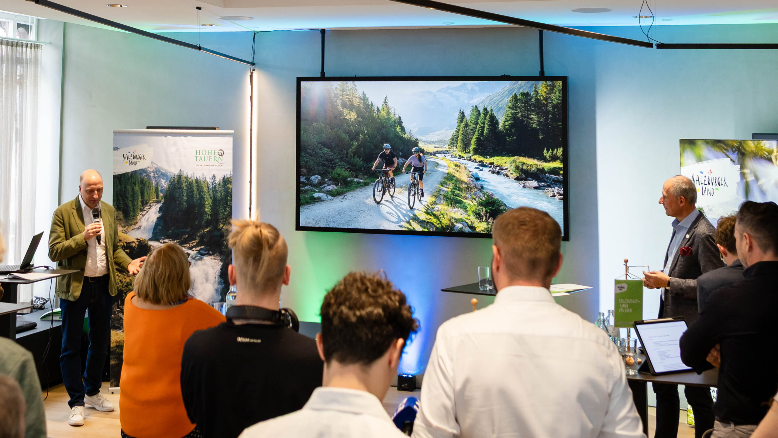 Ein Redner präsentiert dem Publikum auf einer großen Leinwand zwei Radfahrer, die an einem Gebirgsfluss entlang fahren.